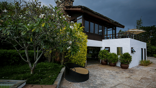 The Serai Bandipur resort exterior with a rustic stone chimney, dark wood upper structure, white walls, potted plants and a landscaped courtyard.