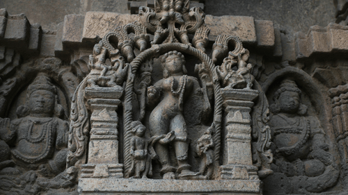 A close up of a stone carving of a dancing lady in Hoysaleswara Temple, Halebidu with other stone carvings of deities next to it.