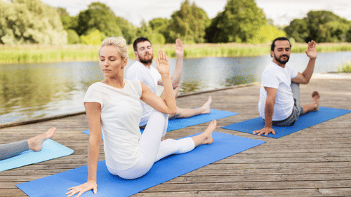 Group of 3 people practicing seated yoga twists on yoga mats on a wooden deck by a lake, surrounded by trees at YO1 Longevity and Health Resorts, Catskills