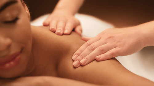 A person lying on their stomach with their eyes closed and a couple of hands massaging their shoulder
