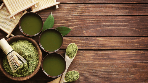 Matcha tea is presented in three cups and a wooden bowl of powder, with a bamboo whisk and spoons, on a wooden surface.