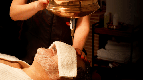 A person lies with eyes covered by a towel, receiving a steady stream of liquid on their forehead from a brass vessel at YO1 Longevity & Health Resorts