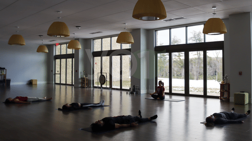 A spacious, well-lit yoga studio with several individuals lying on mats, and one person meditating near large windows, under hanging yellow dome lights at YO1 Longevity & Health Resorts.