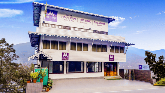 Mount Himalayan Hotel & Spa in Gangtok featuring a multi-level white building with wooden accents, large windows, and panoramic Himalayan mountain views under a clear blue sky.