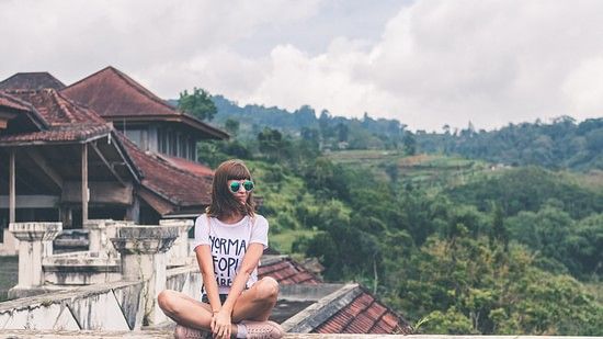 A girl sitting on a ledge with a cottage behind her and forest cover in the background