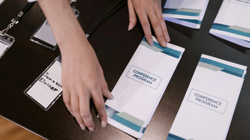 Hands arranging conference programmes and name badges on a dark table at a professional event registration desk.