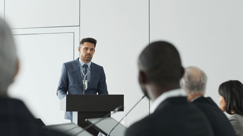 A man in a blue suit stands at a lectern, delivering a speech to a seated audience at a professional conference.