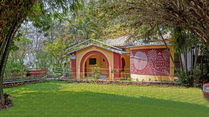 Exterior view of a resort reception building painted in red and yellow, featuring a arched entrance and traditional Warli art mural on the wall, surrounded by lush green lawn and tall trees on a sunny day.