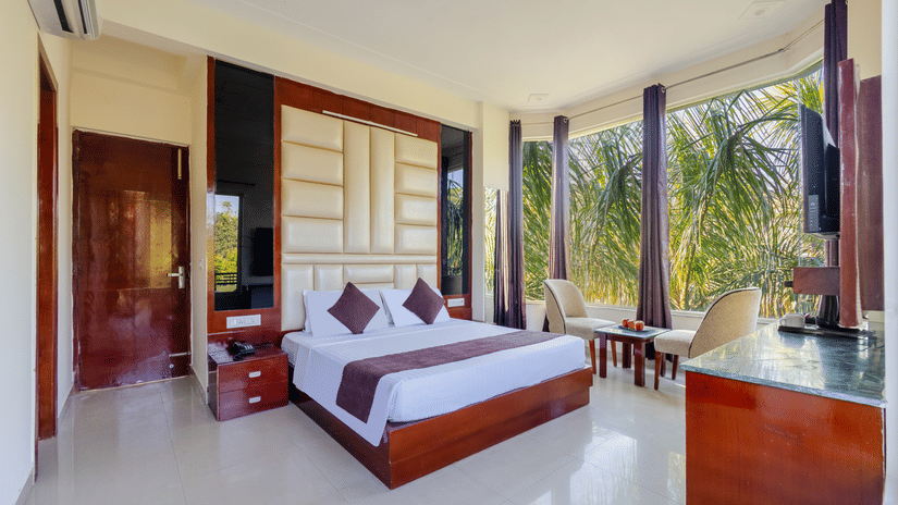 A spacious hotel bedroom featuring large windows with mountain views and a high-set cushioned headboard - Nature Trails Rishikesh