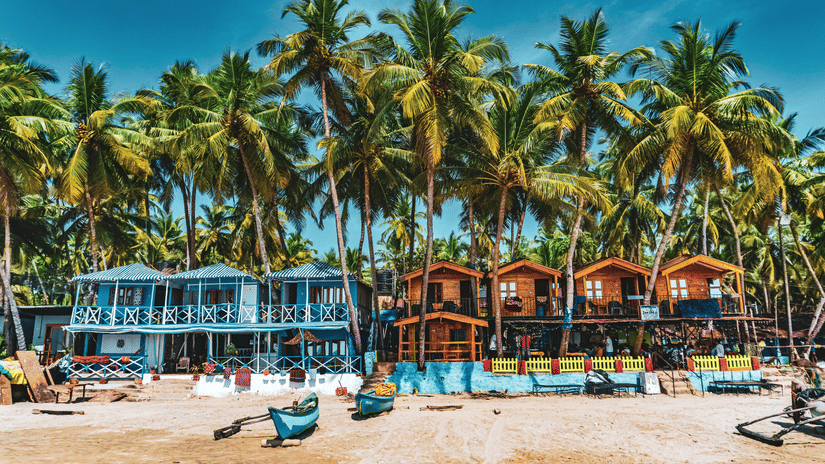 Colourful beach resort with palm trees and blue wooden cabanas on a sandy shore.