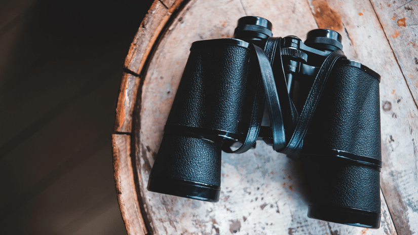 Black binoculars placed on a round wooden surface, with textured tabletop and dark background visible around the object.