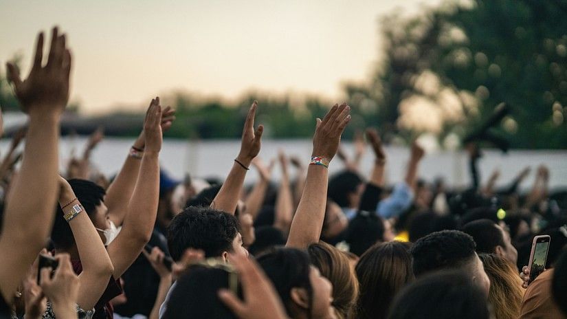 A lively crowd at an outdoor concert with raised hands, wristbands, and people capturing the moment on their phones.