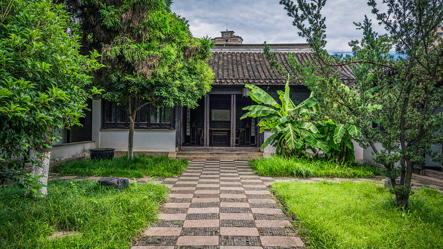 A traditional courtyard with a tiled-roof building, lush greenery, and a patterned stone pathway in the foreground.