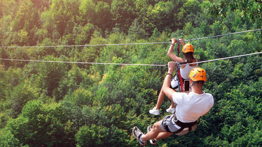 A man and a woman ziplining beside each other during daytime over a lush forest.
