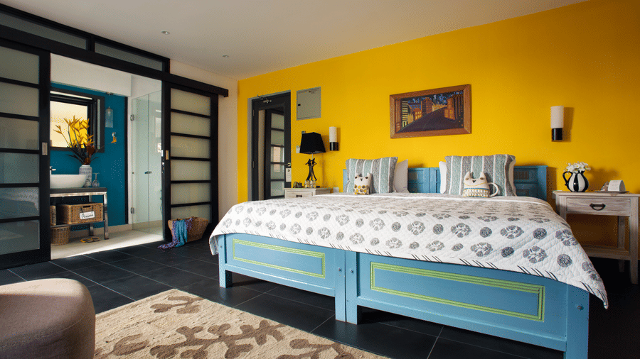 Bright Owl and the Pussycat Hotel in Galle room with a turquoise bed, yellow accent wall, patterned bedding, modern furnishings, and an open bathroom area separated by glass doors.