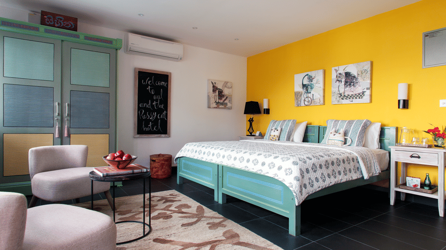 Stylish guest room at Owl and the Pussycat Hotel in Galle featuring a turquoise bed, yellow accent wall, patterned rug, pastel armchairs, and eclectic decor.