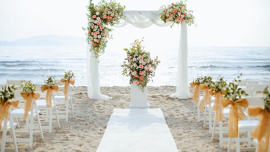 Beachside wedding set up featuring a stage decorated with flowers and fabric pieces. At the centre there is a aisle and on either side there are chairs decorated for guests.