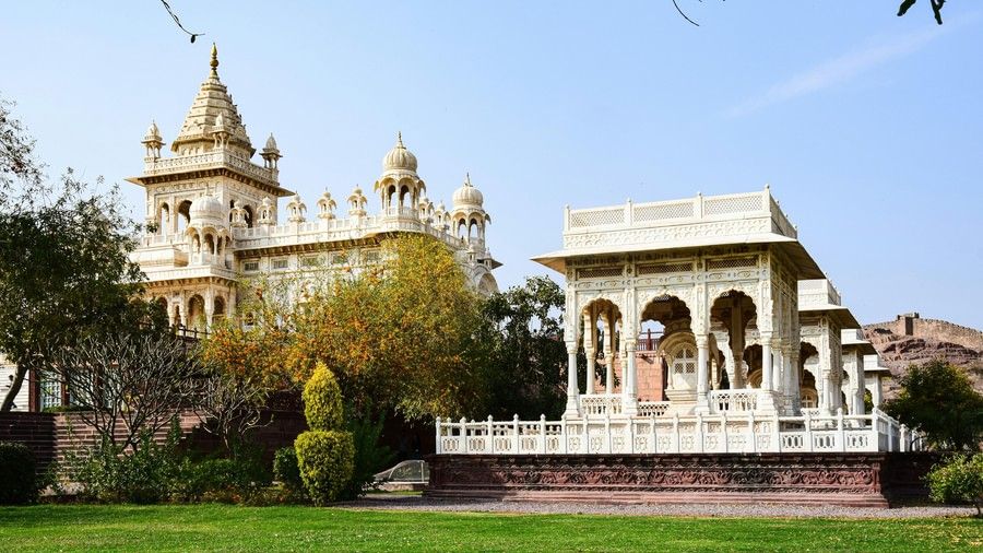 A palace with multiple domes and spires stands next to an open structure with pillars, all surrounded by green grass and trees.