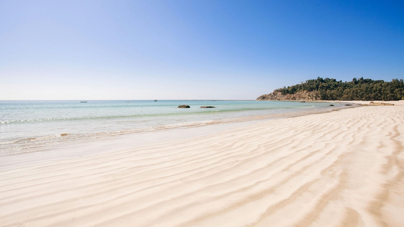 Wide, empty sandy beach with clear blue skies and a distant green hill under bright sunlight.