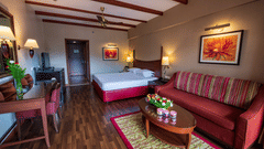 A room with a bed, maroon sofa, a table & a work desk & chair, wall lights and wall art at one of the finest rooms in Kodaikanal for family at The Clarton.