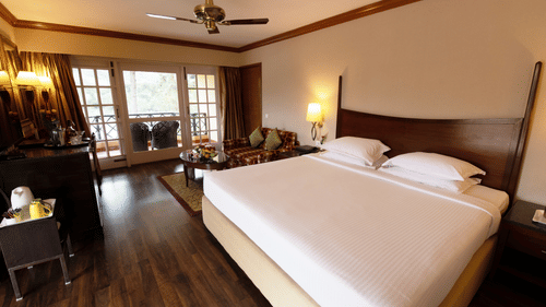 A room with a double bed, wooden furniture & lamps & a large ceiling to floor window with a seating space. Find your ideal Kodaikanal room-booking available now at The Clarton.