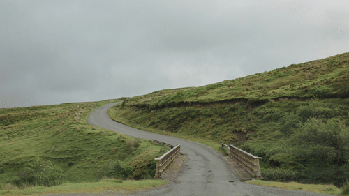 A winding road passing through green hills under a cloudy sky.