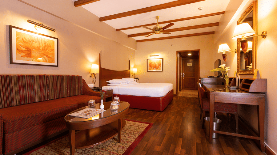 A bedroom at The Carlton in Kodaikanal featuring a double size bed, a work desk and chair, TV screen, a sofa and centre table.