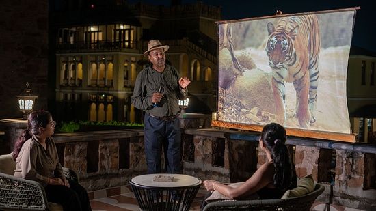 A man stands next to a projector, displaying an image of a tiger and explaining something and two people sitting on chair and listening to him