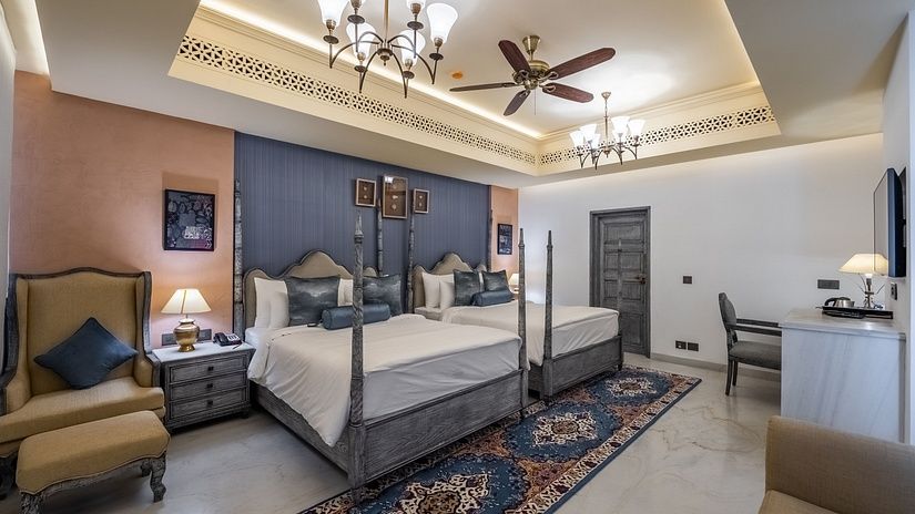 Elegant hotel room with two four-poster beds, vintage-style armchairs, a chandelier, and a writing desk.
