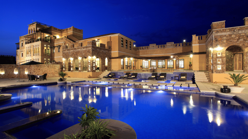 Luxury poolside view of Juna Mahal, Ranthambore at night, featuring illuminated heritage style architecture, spacious outdoor seating, and a large swimming pool reflecting the resort's lights.