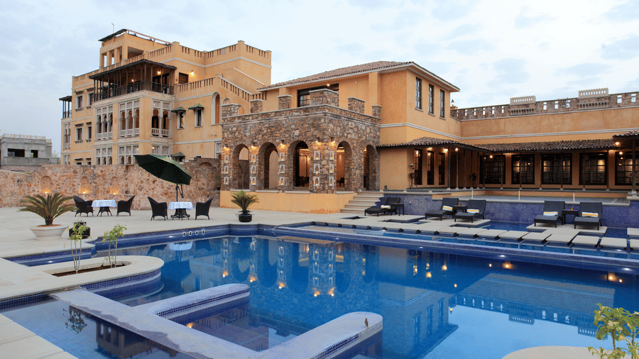 Outdoor swimming pool at Juna Mahal, Ranthambore, surrounded by heritage inspired architecture, stone archways, poolside loungers, and open seating areas overlooking the resort.