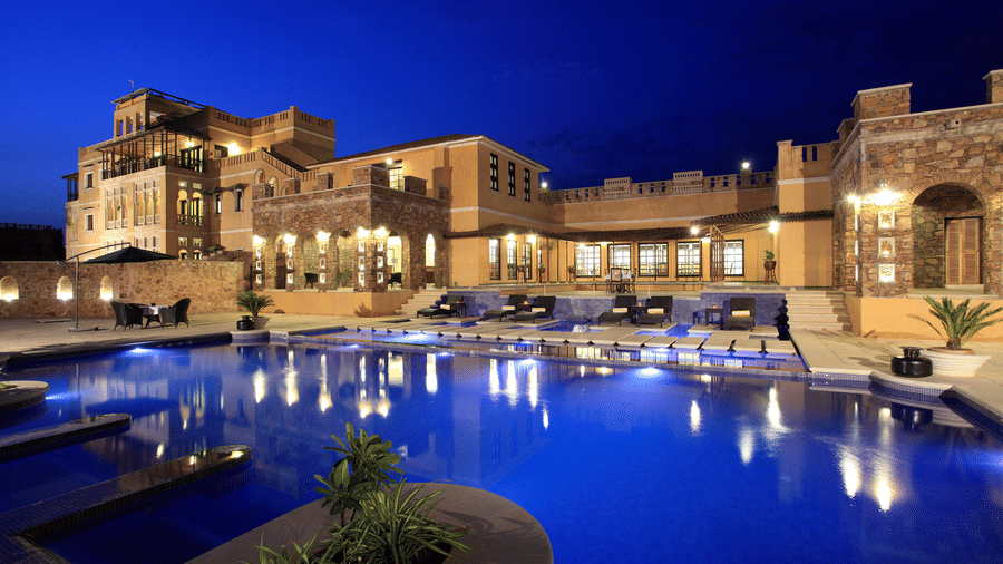 Luxury poolside view of Juna Mahal, Ranthambore at night, featuring illuminated heritage style architecture, spacious outdoor seating, and a large swimming pool reflecting the resort's lights.