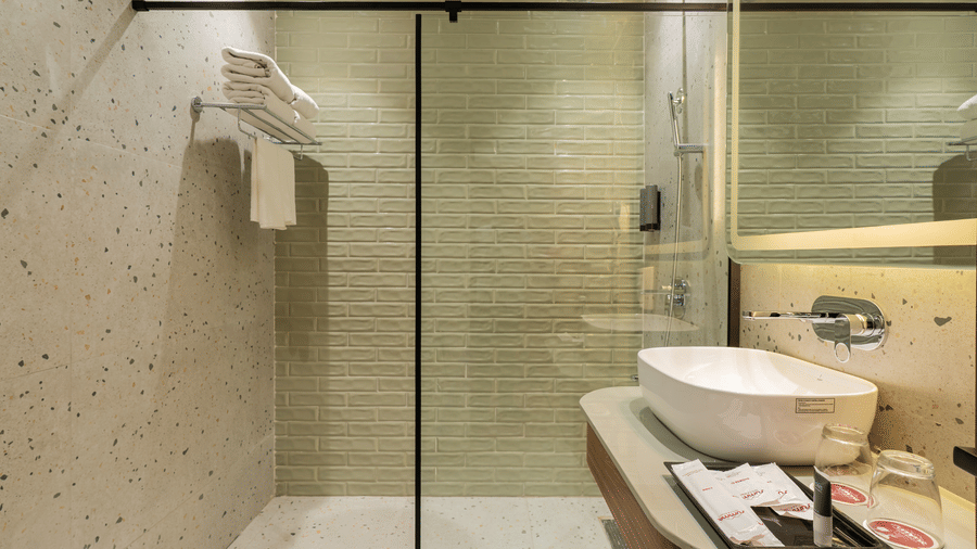 A bathroom with a walk-in shower behind a glass partition, a big mirror with a wash basin, toiletries on a counter top, and fresh towels on a wall shelf | Summit by the Ganges Beach Resort & Spa, Rishikesh