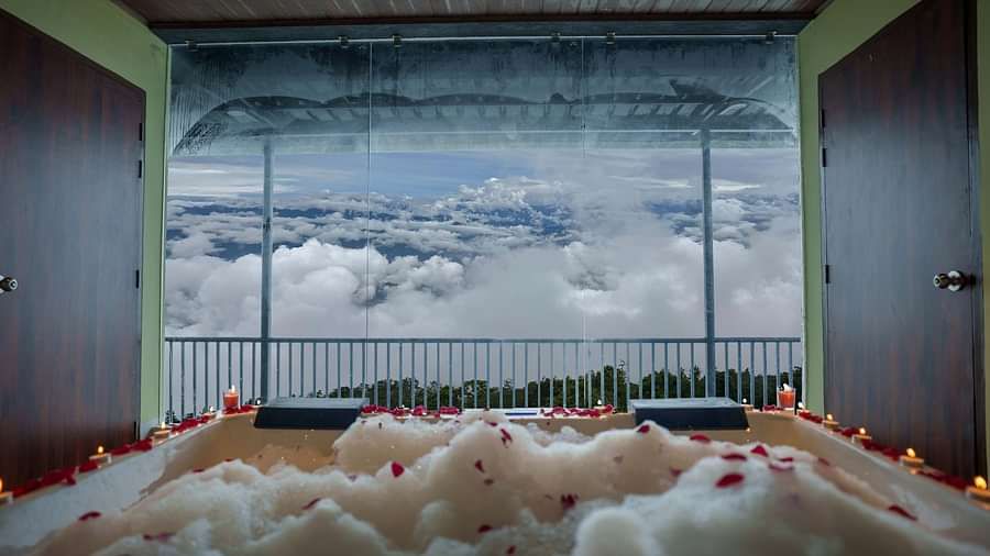 A jacuzzi with rose petals scattered, leading to a balcony overlooking a cloudy sky and distant hills, suggesting a romantic setup at Summit Enigma Resort and Spa.