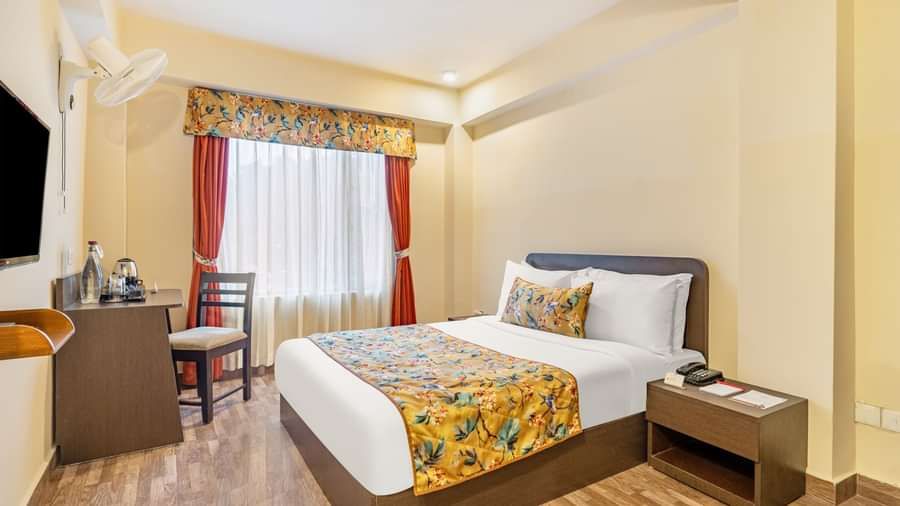 Well-lit hotel room with a double bed featuring floral bedding, a window with red curtains, a small work desk, wall-mounted TV, and wooden flooring - Summit Namnang Courtyard & Spa, Gangtok
