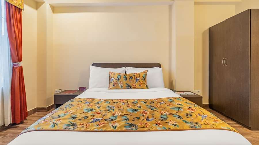 Hotel room with a neatly made double bed, floral yellow bedding, matching pillow, bedside tables, wardrobe, and a window with curtains - Summit Namnang Courtyard & Spa, Gangtok