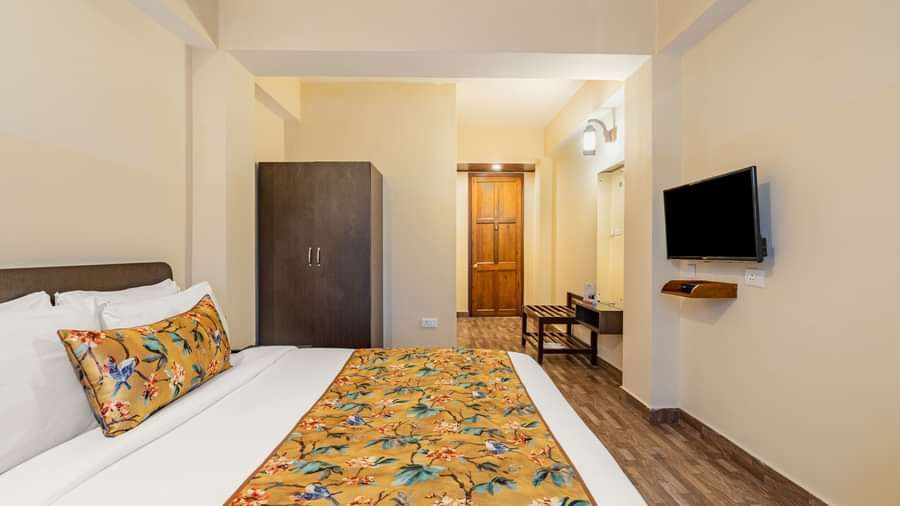 Spacious hotel room with a double bed, floral yellow bedding, wardrobe, wall-mounted TV, wooden flooring, and seating area near the entrance - Summit Namnang Courtyard & Spa, Gangtok