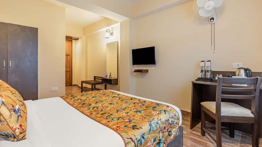 Hotel room with a double bed, floral bedding, wardrobe, wall-mounted TV, study desk with water bottles and kettle, wooden flooring, and ceiling fan - Summit Namnang Courtyard & Spa, Gangtok