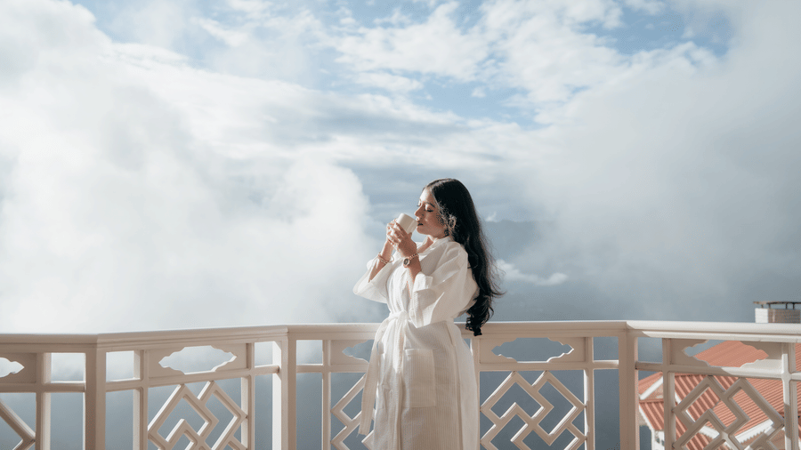 A woman wearong a bathrobe standing in a balcony sipping a hot beverage.