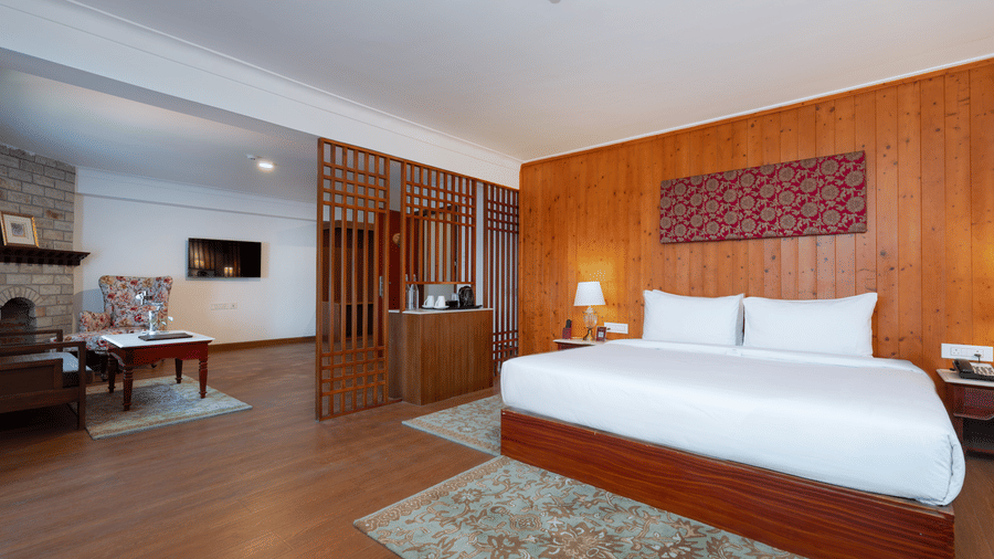 A bedroom at Summit Norbu Ghang - The Signature Collection, Gangtok, with a king size bed, bedside lamps, wooden flooring, sofa, wall-mounted TV, and a table.