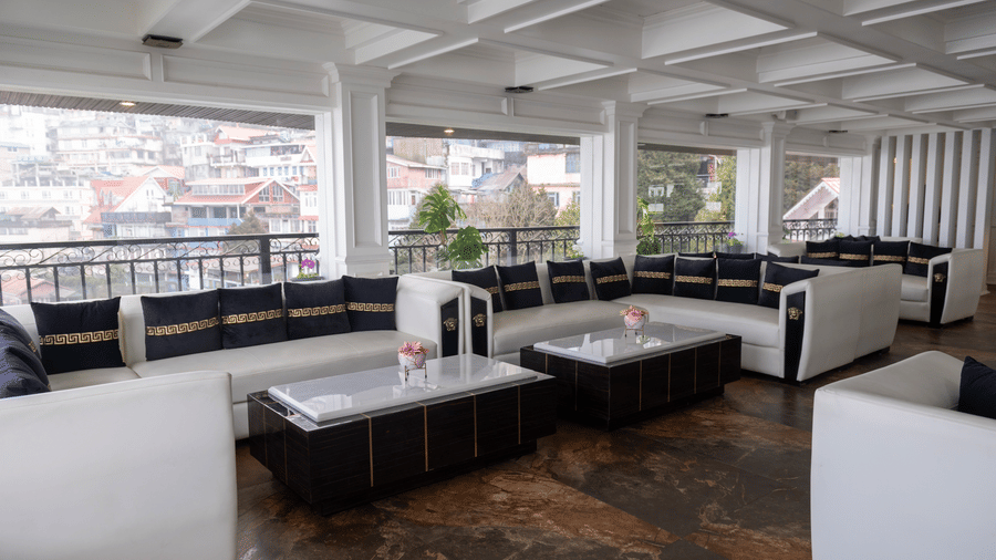 Lounge area at The West Gate Posada By Summit, Darjeeling, featuring sets of sofas arranged around tables facing windows looking out over hills.