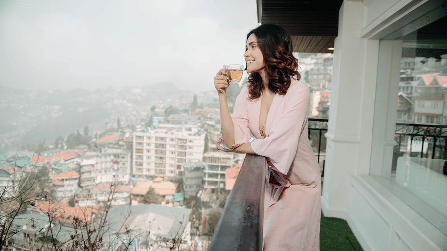 A person standing on a balcony at The West Gate Posada By Summit, Darjeeling, holding a cup and looking out over hills and buildings.