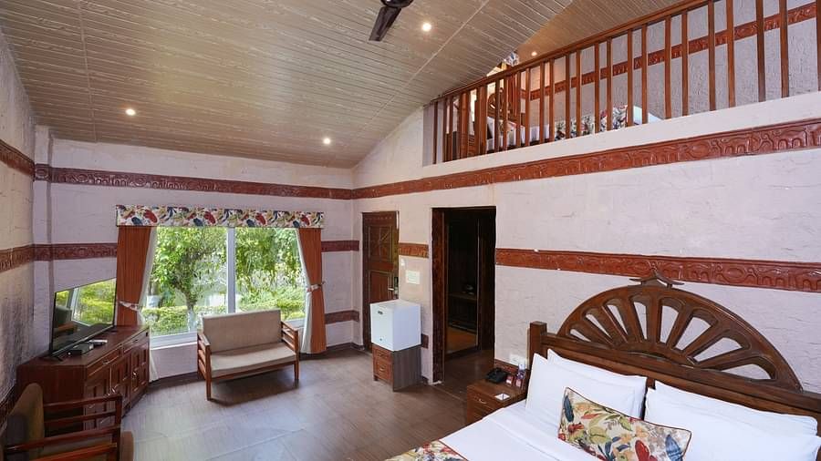 Duplex ethnic villa showcasing a spacious bedroom furnished with wooden furniture, television, and large windows adorned with curtains at Summit Green Lake Tea Resort & Spa, Kaziranga