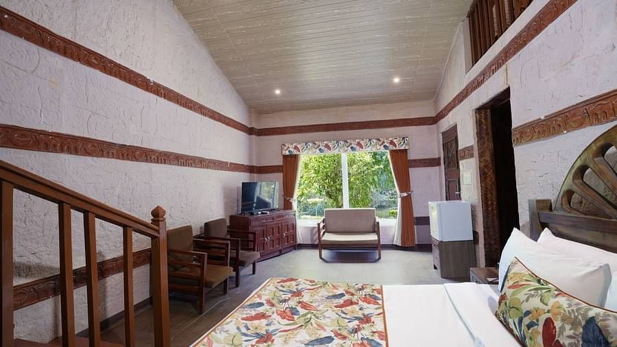 Duplex ethnic villa showcasing a spacious bedroom furnished with wooden furniture, television, and large windows at Summit Green Lake Tea Resort & Spa, Kaziranga