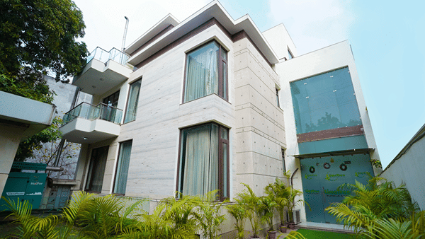 Entrance with a stone facade, branded glass doors with wreaths, potted areca palms, a standby generator, and artificial grass.