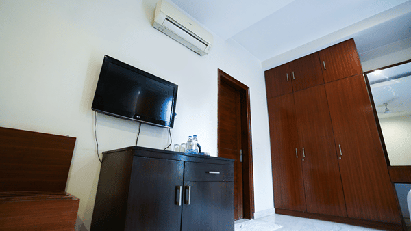Guest room detail with a wall-mounted LG TV, a split air conditioner, a dark minibar with water bottles, and a wood wardrobe.