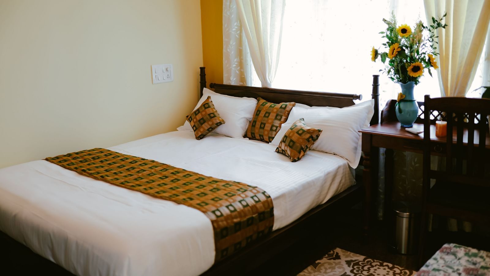 A bedroom with a wooden bed with white linens and patterned throw pillows and a throw blanket, next to a small table | Adhyaya Heritage Reverbed