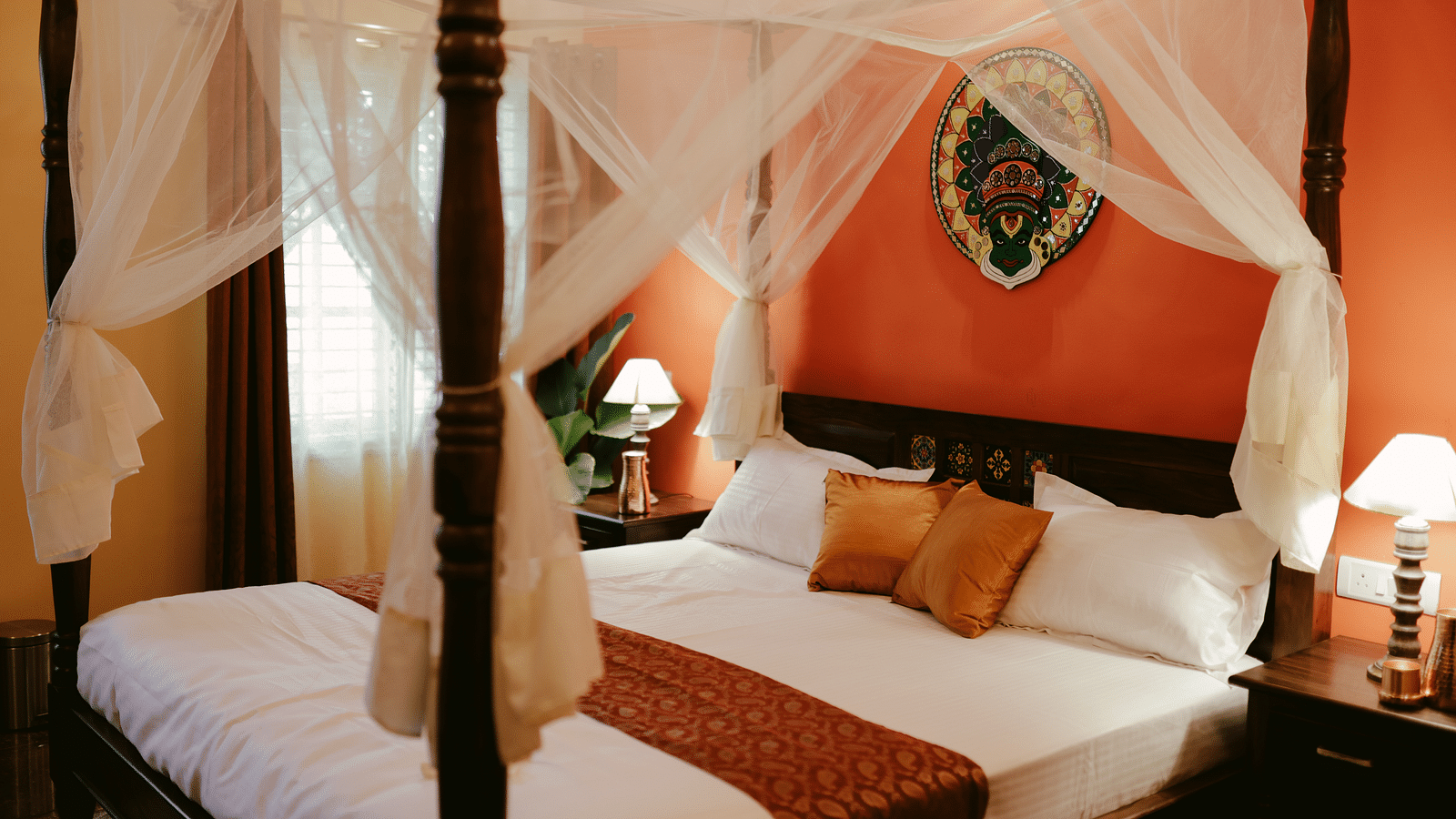 A four-poster bed with white drapery and a decorative headboard in a room with an orange accent wall | Adhyaya Heritage Reverbed