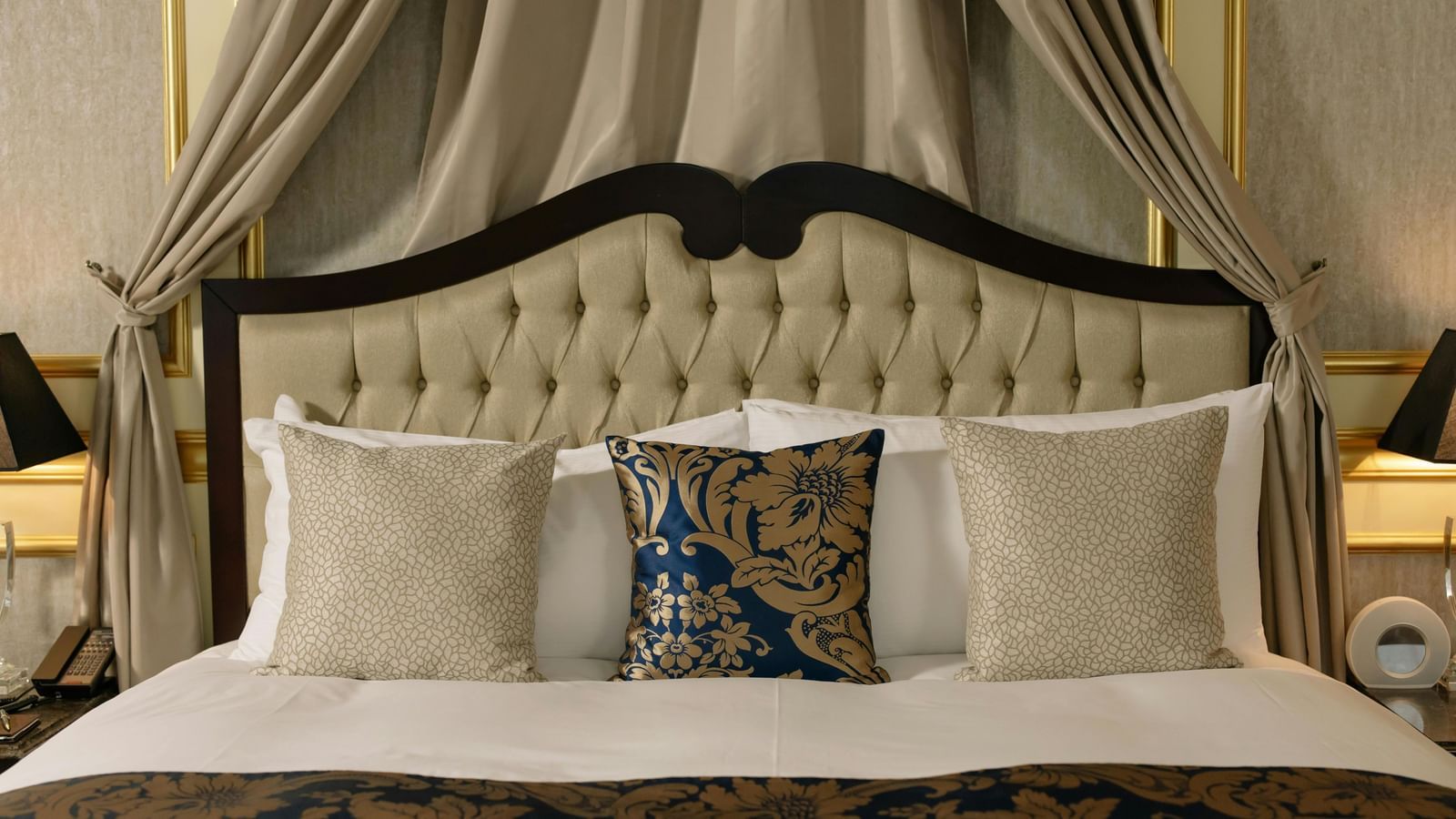 A luxurious hotel room features a large bed with a canopy and elegant bedding, flanked by bedside tables with lamps.