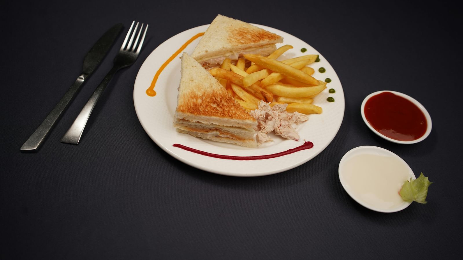 A plate of sandwich, is served with dipping sauces and a lime wedge on a dark surface.